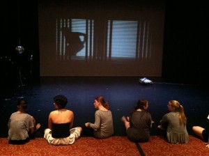 Dancers sit in front of the stage as the audience takes their seat.